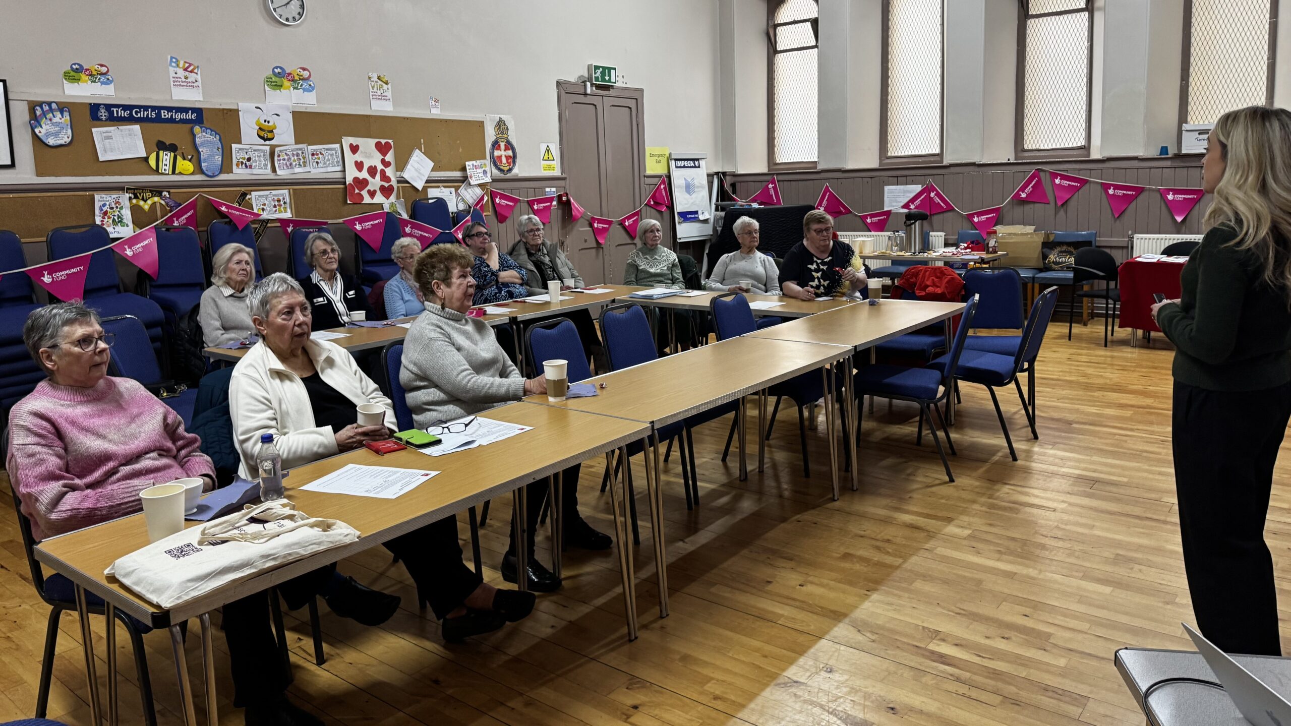 Older adults taking part in a digital safety workshop in a community hall, with a presenter speaking at the front of the room.
