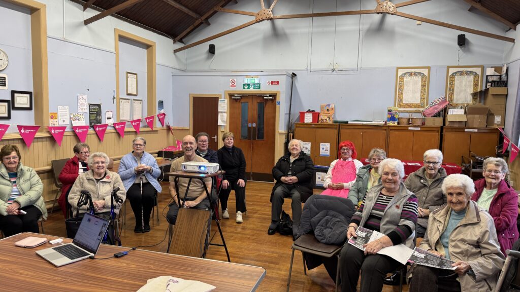 A group of older adults smiling during a digital safety workshop in a community hall, with a laptop and projector set up for the session.